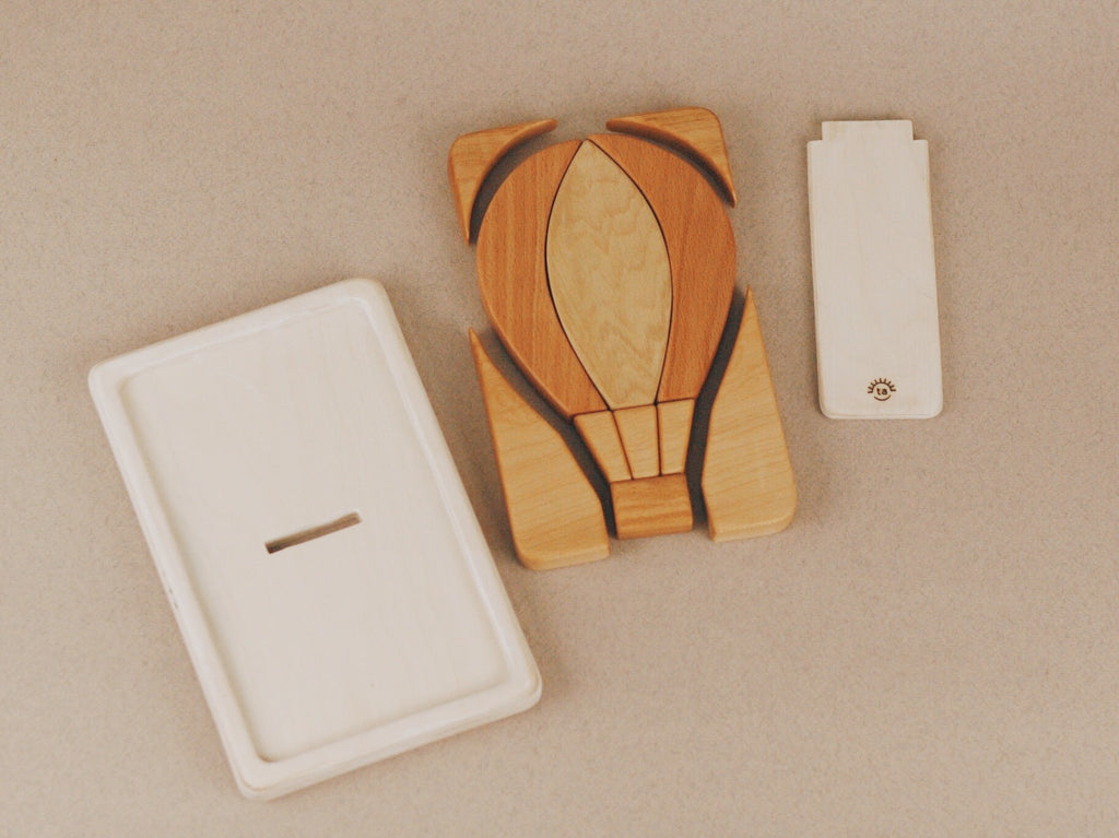 The Handmade Hot Air Balloon Puzzle features wood pieces treated with linseed oil. It's accompanied by a simple rectangular wooden box with a lid and detachable kickstand.