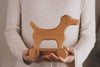 A person in a beige sweater holds the Handmade Wooden Dog Pull Toy, crafted from smooth maple and beech wood and featuring simple carved details along with four wheels.