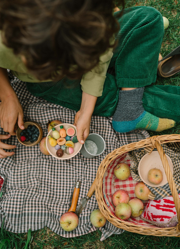 Wearing green corduroy pants, a person enjoys a picnic with a bowl of candies resembling the Grapat Play Set - Fruits. On the checkered cloth are apples, berries, wooden fruits, a basket, and utensils—perfect for open-ended play.