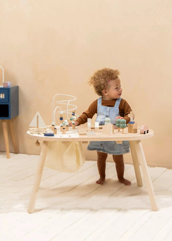 A toddler with curly hair playing with wooden toys on a Wooden Activity Table in a room with soft beige walls and floor.
