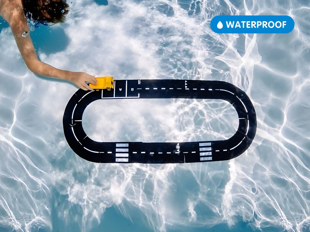 A hand pushes the yellow WaytoPlay Medium Flexible Toy Road Set - Expressway on a black racetrack mat floating in a pool, sunlight shimmering on the water. Made from unbreakable child-safe material, with a "WATERPROOF" label in the top right corner.