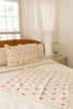 A cozy, light-filled bedroom with a wooden bed frame, cream bedding, and decorative pillows and throw in simple brown geometric patterns. An Organic Cotton Layered Gauze Pillowcase - Teddy adds soft texture, beside a lamp near sheer-curtained windows.