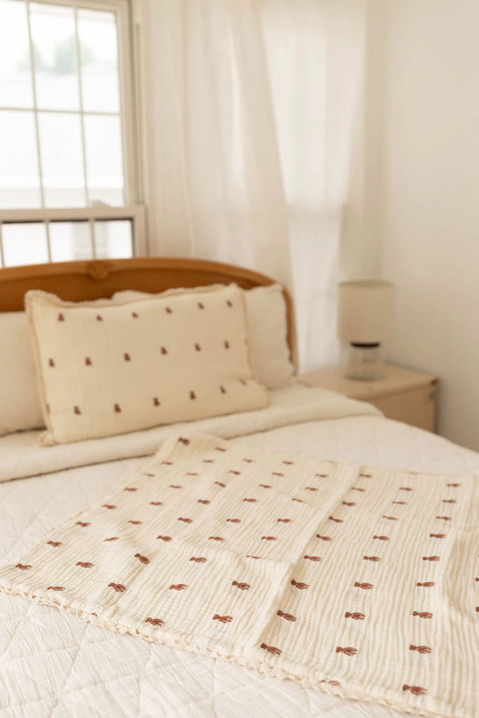 A cozy, light-filled bedroom with a wooden bed frame, cream bedding, and decorative pillows and throw in simple brown geometric patterns. An Organic Cotton Layered Gauze Pillowcase - Teddy adds soft texture, beside a lamp near sheer-curtained windows.