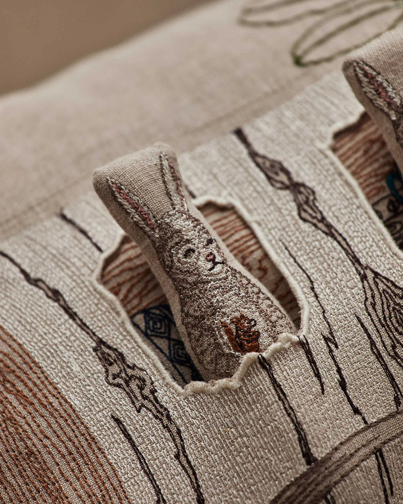 A close-up of the Coral & Tusk Aspen Log Bunnies Pocket Pillow shows embroidered bunnies peeking through a cut-out window, surrounded by detailed stitching, textured patterns, and neutral-toned linen fabric.