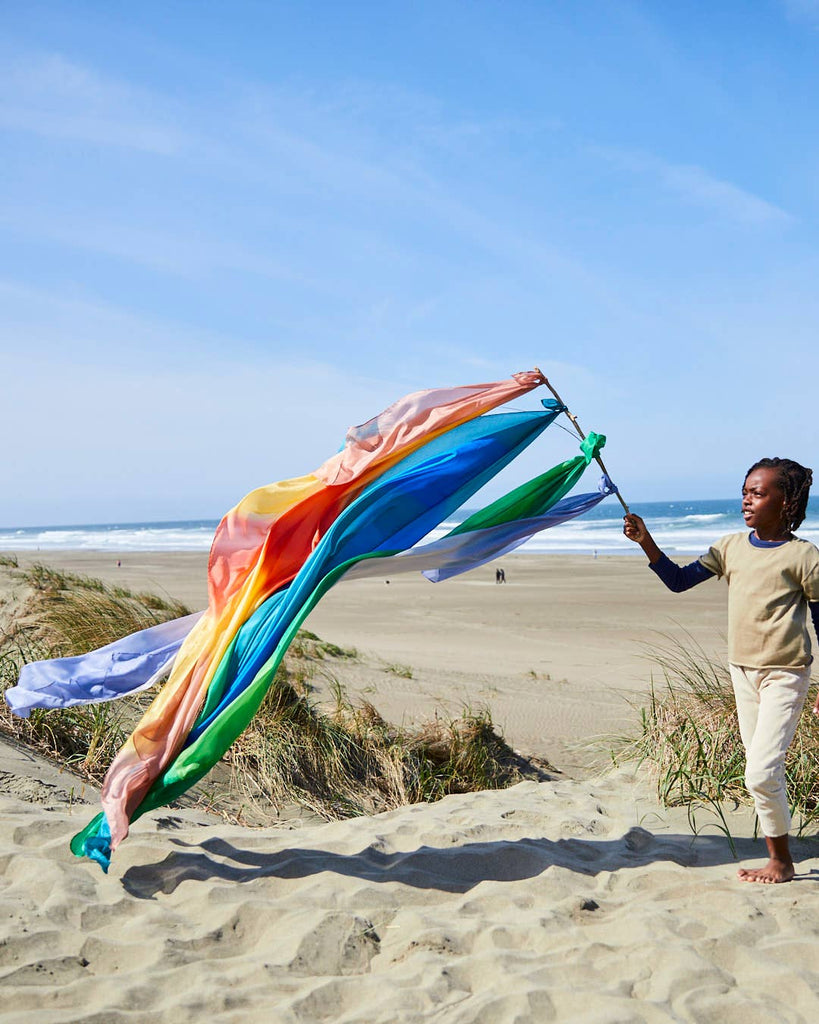 A child stands on a sandy beach, gripping a long pole adorned with Sarah's Silk Giant Ocean Playsilk billowing in the breeze. Immersed in imaginative play, the vibrant fabric sways gracefully against the tranquil backdrop of ocean and sky, while a few figures appear faintly in the distance.