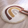 A child’s bare legs and feet rest on the edge of the Bohemian Rainbow Children's Rug - Final Sale, a round rug featuring yellow, brown, and gray boho arches on a wooden floor.