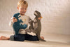 A young child sits on the floor, smiling and holding a Steiff Thaisen Tyrannosaurus "T-Rex" Dinosaur Plush Toy (14 inches) and a blue dino plush, with a white brick wall in the background.