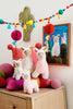 A Steiff Leandro Llama Plush Toy, 7 inches, sits on a dresser adorned with colorful felt balls and pillows. Above, llama photos, cactus wall art, and a garland with pom-poms and paper lanterns add cheerful accents.