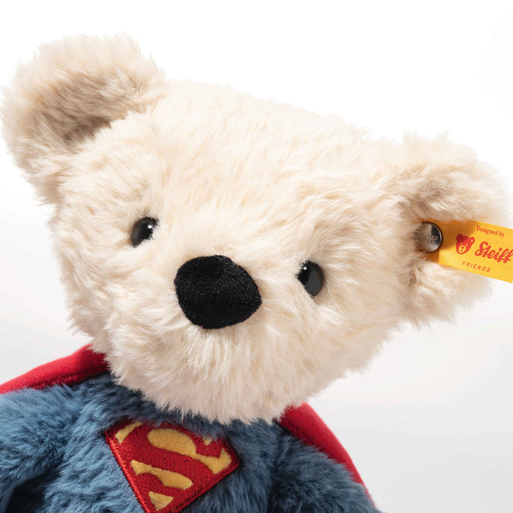 The Steiff Superman Teddy Bear, 12 Inches, is a soft beige plush bear wearing a blue outfit with a red cape and the iconic Superman "S" emblem. A yellow tag in its ear makes this adorable DC collectible perfect for fans.