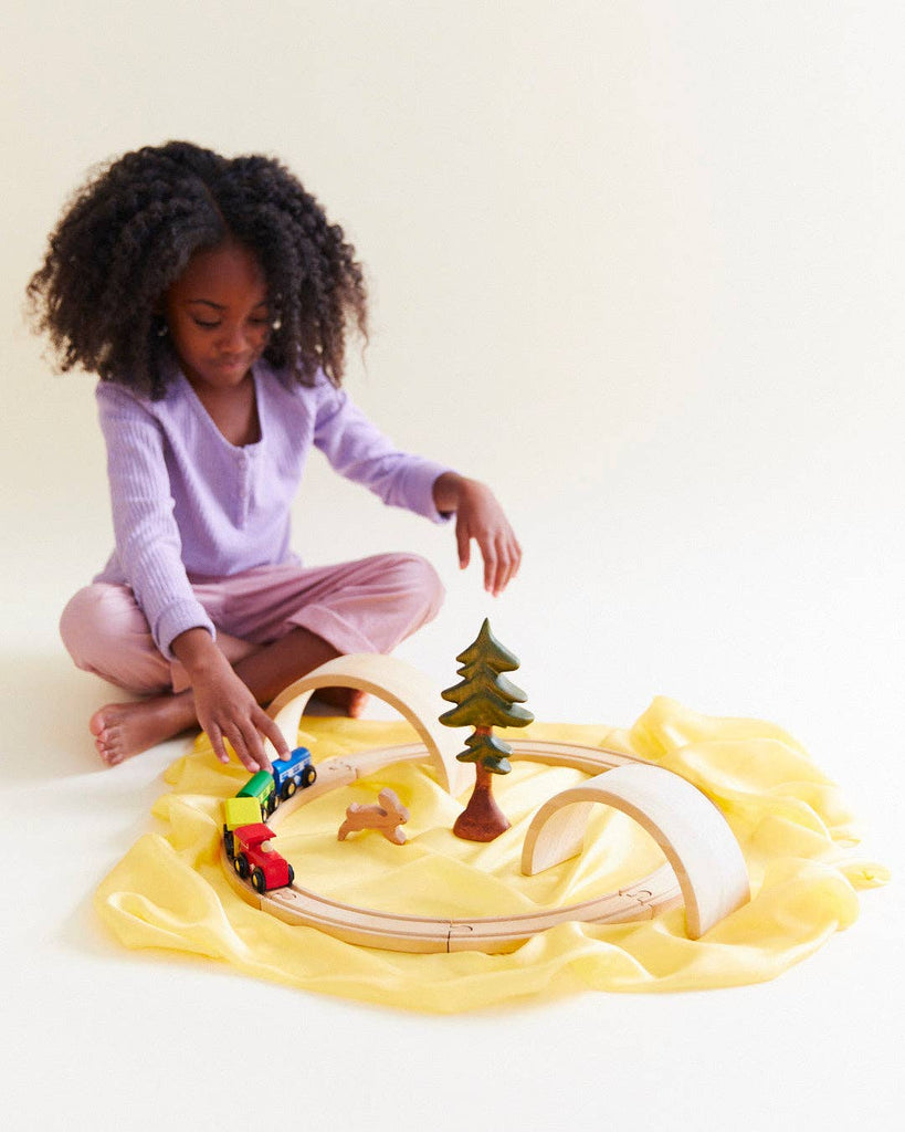 A young girl with curly hair, in a light purple top and pink pants, sits on a Sarah's Silks Solid Color Playsilk - Sunshine, enjoying open-ended play with a wooden train set, curved tracks, arches, and a toy tree.