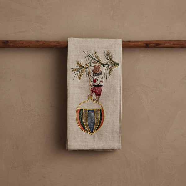 The Coral & Tusk Deck the Halls Bear Tea Towel in beige linen features an embroidered bear in red shorts on a colorful ball with an olive branch—perfect as festive holiday kitchen decor or a Christmas tea towel.