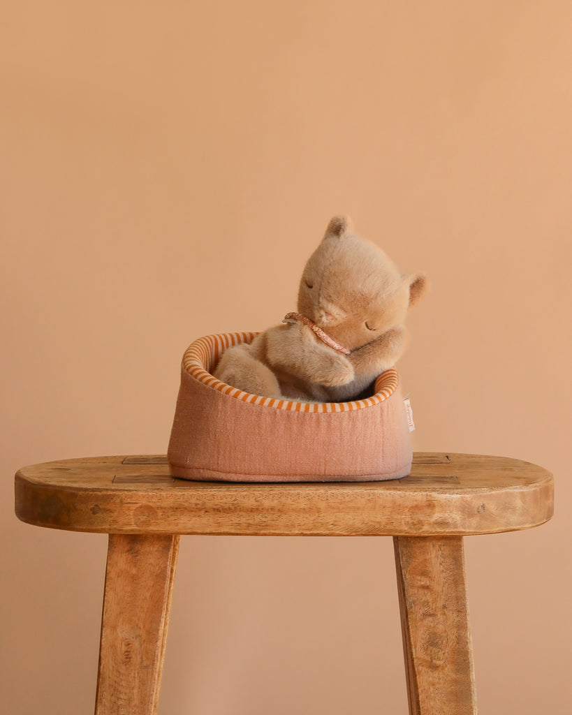 The Maileg Small Sleeping Kitten in Basket features a plush kitten with closed eyes resting in a round, peach-colored basket on a wooden stool, set against a beige background—an ideal match for Maileg accessories.