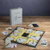 The Clue Vintage Bookshelf Edition is displayed on a wooden table with colorful pieces, cards, notepads, pencils, and its distinctive book-shaped box standing upright in the background.