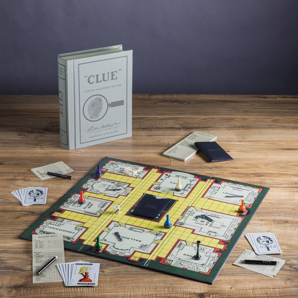 The Clue Vintage Bookshelf Edition is displayed on a wooden table with colorful pieces, cards, notepads, pencils, and its distinctive book-shaped box standing upright in the background.