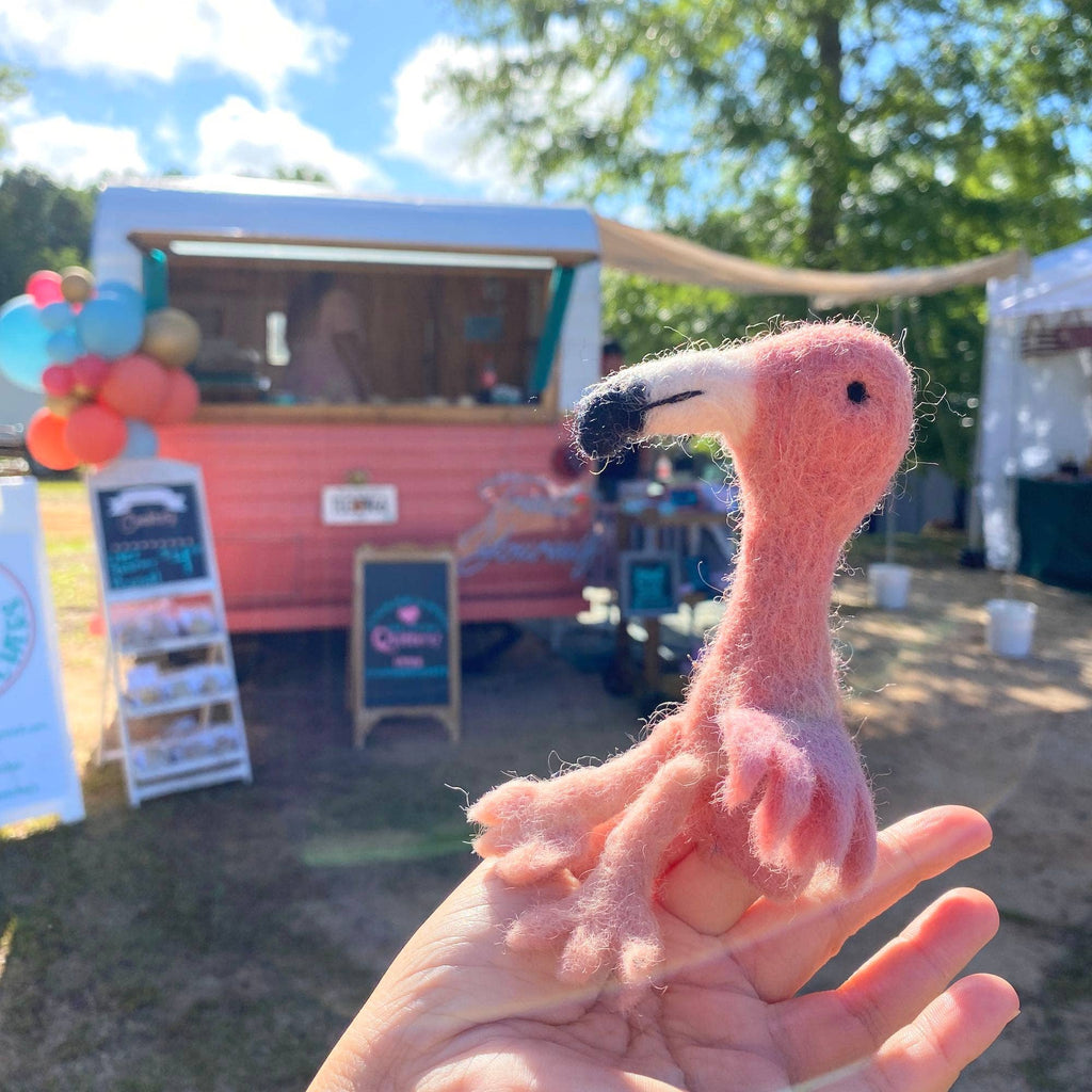 A hand holds a Flamingo Finger Puppet in front of a pink food truck decorated with balloons. Trees, a chalkboard sign, and a sunny sky create an ideal backdrop for imaginative play.