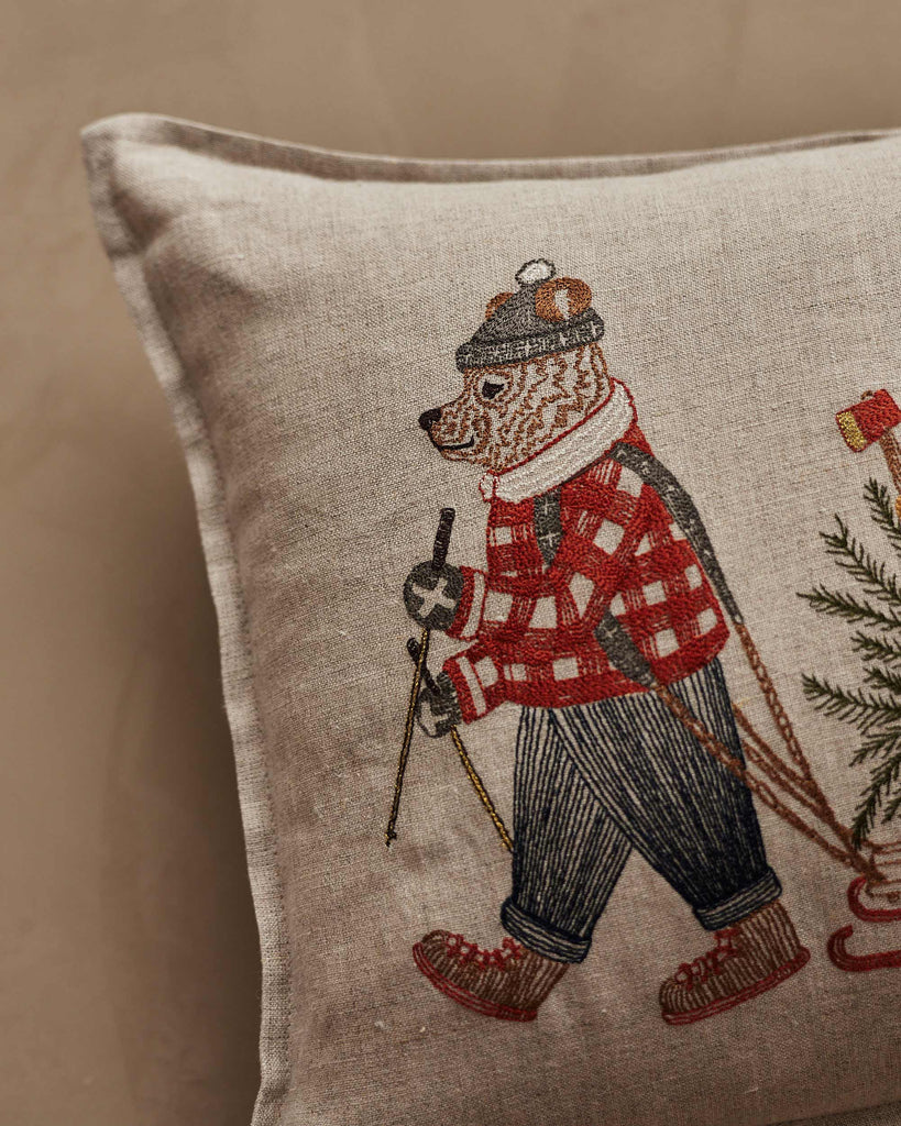 Coral & Tusk Christmas Tree Sled Pillow made of embroidered linen featuring a bear in a plaid shirt and knit hat pulling a wooden sled with a Christmas tree.
