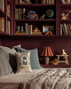 A cozy reading nook with maroon bookshelves filled with books and toys, a cushioned bench decorated with spring pillows—one featuring the Coral & Tusk Kitten Cameo Pillow—a taupe quilt, a small lamp, and wooden animal figurines.