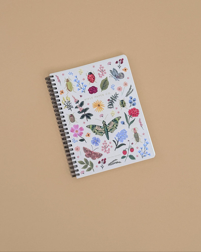 The Curio Spiral Notebook is a softcover spiral notebook with a white cover decorated with colorful flowers, leaves, butterflies, ladybugs, and insects on a beige background. Ideal for jotting notes on its ruled pages.