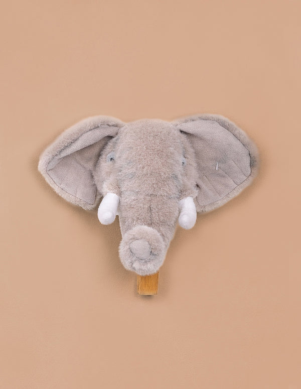 The Coat Hanger - Elephant features a plush gray elephant head with big floppy ears and white tusks on a tan background, serving as both a cute soft toy wall decor and a functional coat hanger.