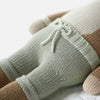 Close-up of the lower half of Cuddle + Kind Elliott the Fawn, a hand knit, hypoallergenic doll wearing light green drawstring shorts, lying on a soft white surface.