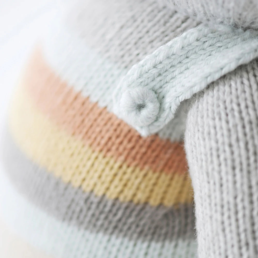 Close-up of Cuddle + Kind Evan the Elephant doll’s hand-knit fabric with pastel gray, orange, yellow, and white stripes, featuring a pale blue knitted strap with a button detail.