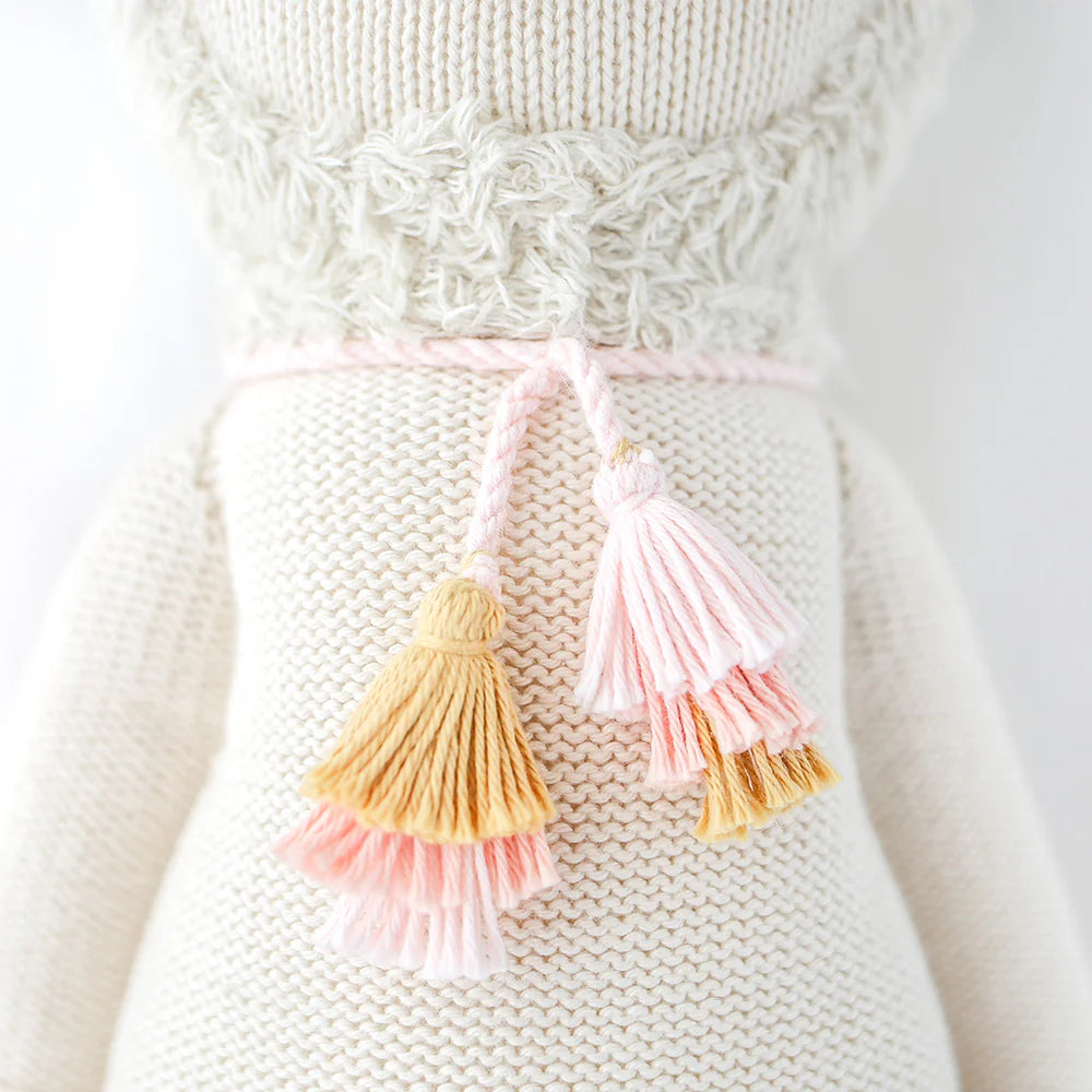 A close-up of the Cuddle + Kind Lola the Llama, a hand-knit doll dressed in a cream outfit with a fluffy textured collar and two pink and yellow tassels on a pink string around its waist.