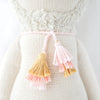 A close-up of the Cuddle + Kind Lola the Llama, a hand-knit doll dressed in a cream outfit with a fluffy textured collar and two pink and yellow tassels on a pink string around its waist.