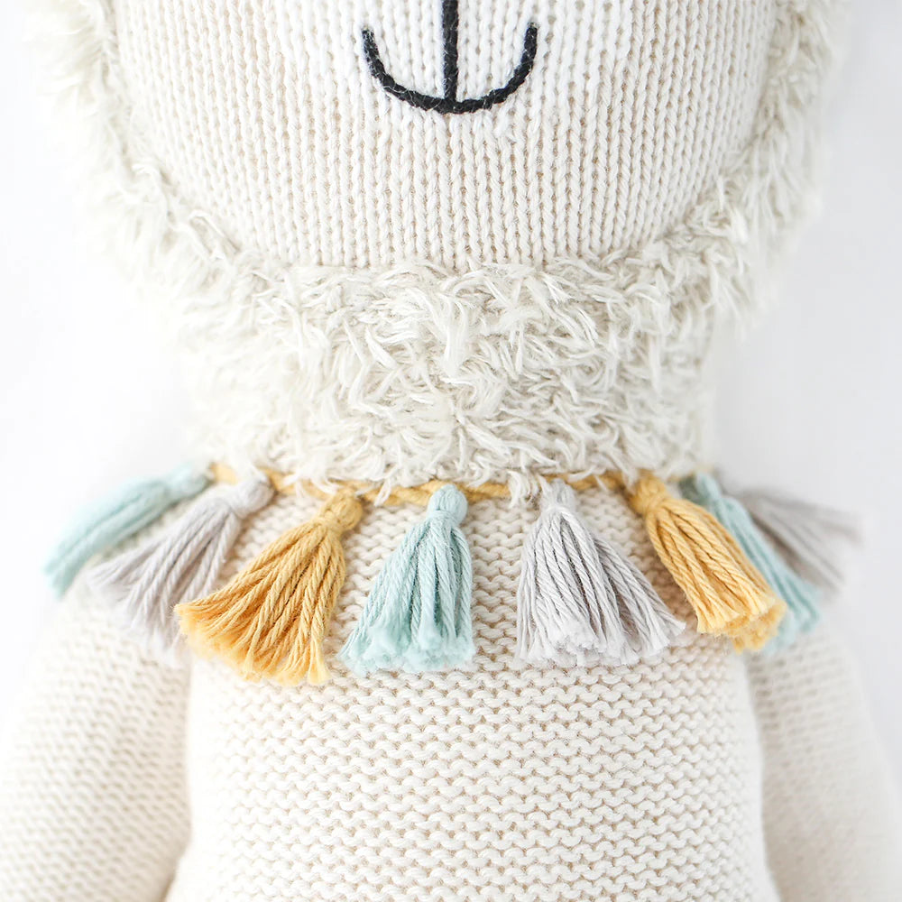 A close-up of Cuddle + Kind Lucas the Llama, a fair trade, hand-knit doll with a stitched black smile and nose, wearing a fringed collar with gray, yellow, and light blue tassels. Made from soft organic cotton.