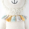 A close-up of Cuddle + Kind Lucas the Llama, a fair trade, hand-knit doll with a stitched black smile and nose, wearing a fringed collar with gray, yellow, and light blue tassels. Made from soft organic cotton.