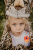 A young blond child with blue eyes wears the Costume - Leopard faux fur hood and a white shirt with an orange and purple flower patch, looking up seriously outdoors against green foliage.