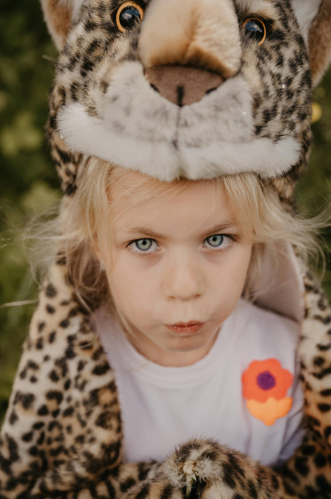 A young blond child with blue eyes wears the Costume - Leopard faux fur hood and a white shirt with an orange and purple flower patch, looking up seriously outdoors against green foliage.