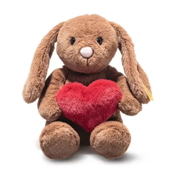 Steiff Hoppie Rabbit – Love is a brown plush bunny with long ears, a pink nose, and black stitched eyes. It sits upright holding a red heart-shaped pillow—a charming gift for Steiff fans. Background is plain white.