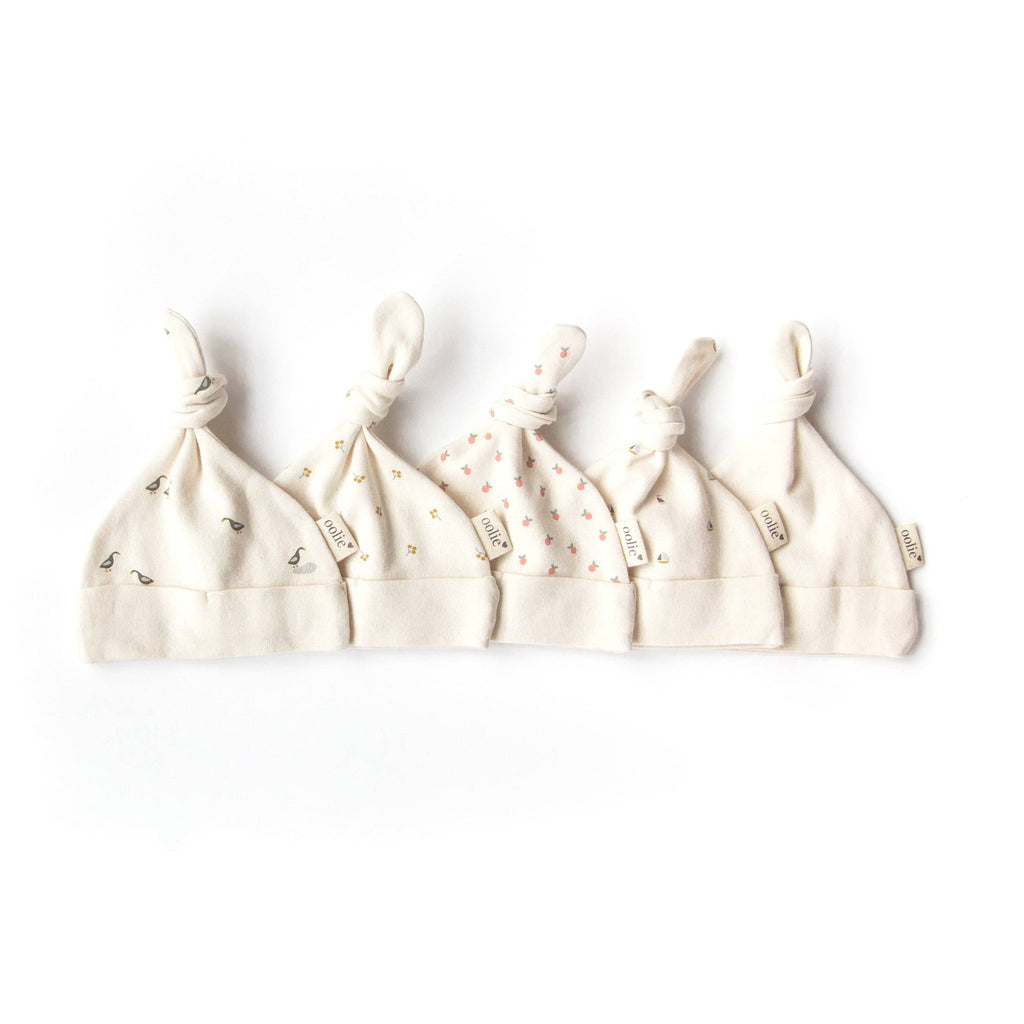 Five cream-colored Organic Baby Hat – Gold Sprig with knotted tops are arranged in a row, each showing a unique minimalist pattern and a fabric tag—crafted from GOTS certified organic cotton, perfect for newborns.