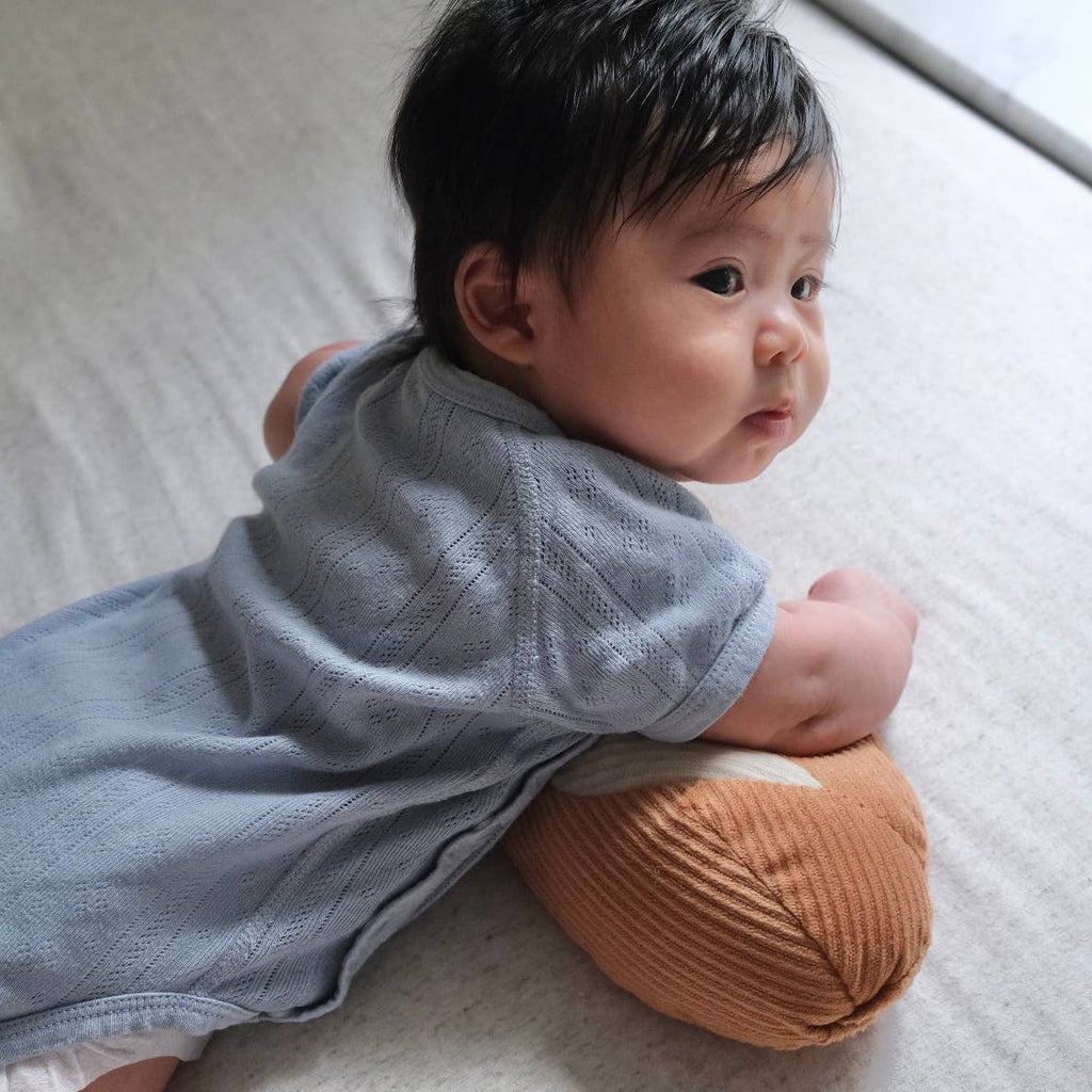 A baby with dark hair, dressed in light blue, lies on their stomach atop a soft surface, propped up on the Tummy Time Baguette pillow and gazing thoughtfully out of the frame.