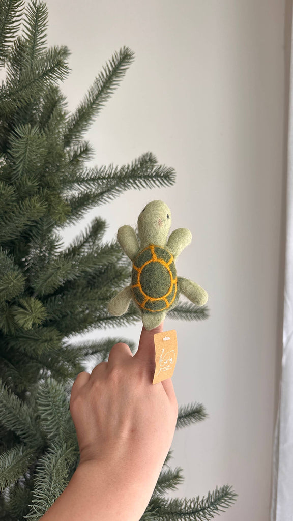 A hand displays the Turtle Finger Puppet—a small, handmade green turtle with an orange shell—in front of a pine tree with sunlight streaming through a window, making it a delightful addition to any Finger Puppet Collection.
