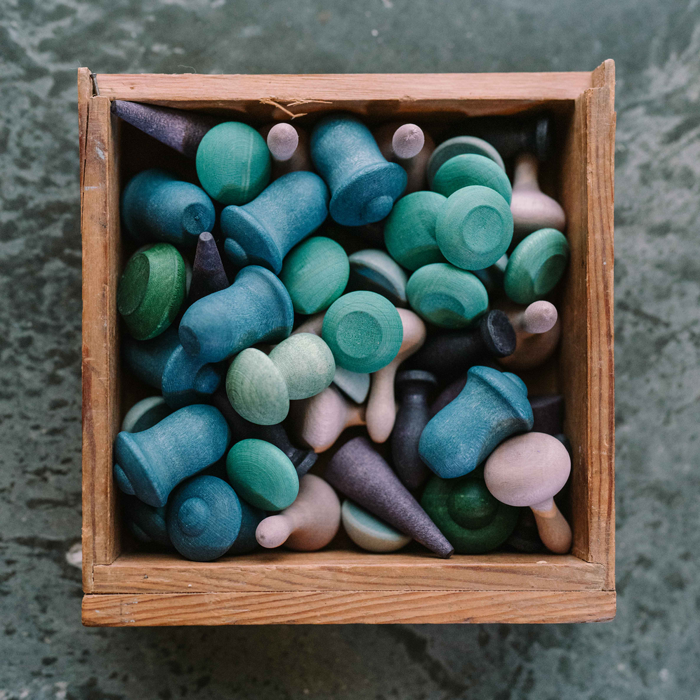 A wooden box filled with Grapat Mandala Mix - Cold toys in green, blue, and purple hues—wooden mandalas and pegs of various shapes—seen from above on a textured surface; perfect sustainable toys for creative play.