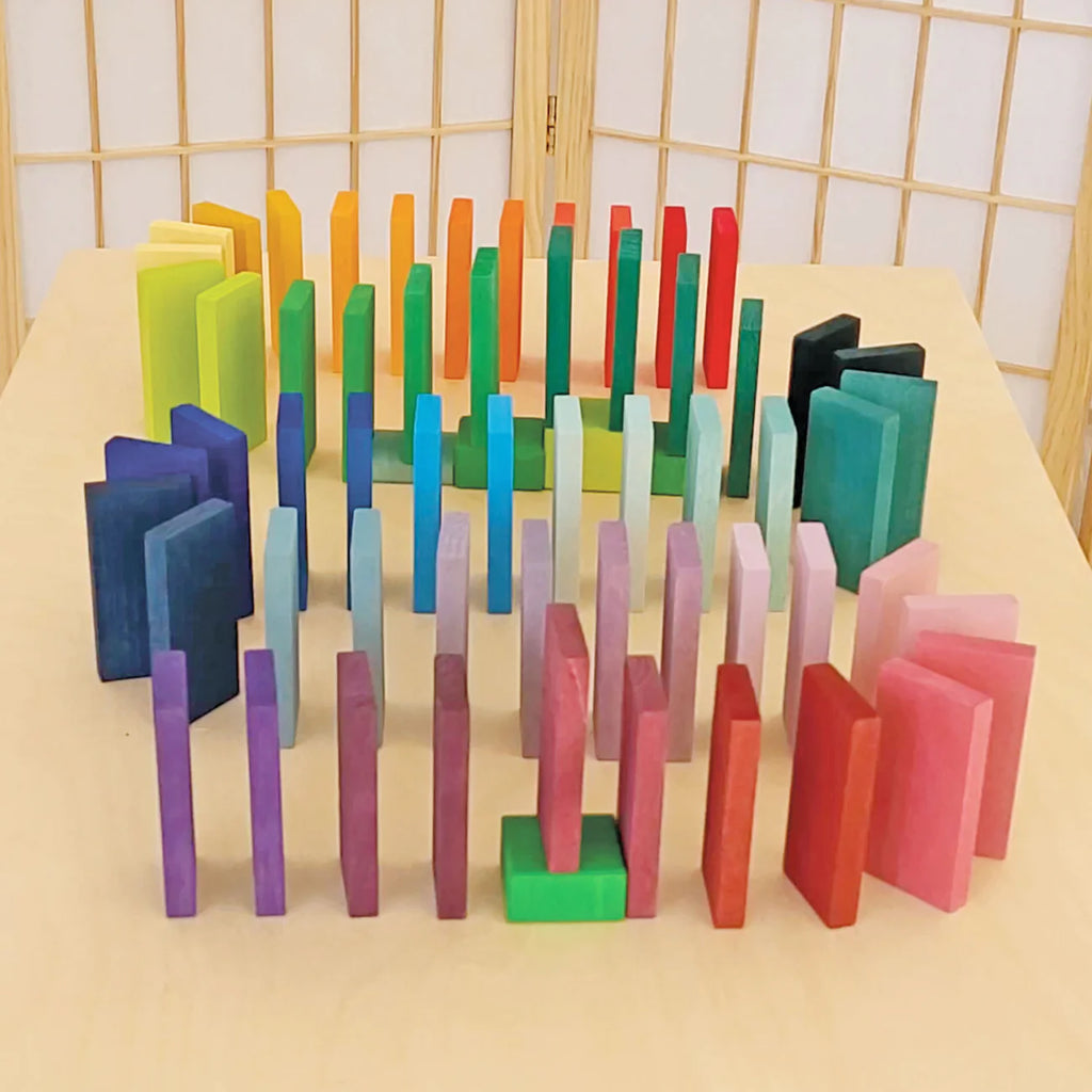 Grimm's Color Charts-Rally features vibrant dominoes arranged upright in concentric circles on a light wooden surface, showcasing a blue, green, yellow, red, and pink gradient with light wood panels as the backdrop.