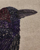 Close-up of the Coral & Tusk Blackbird with Acorn Pillow showcases intricate embroidery of a blackbird on light beige linen, featuring detailed black, purple, and dark red stitching.