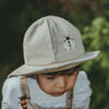 A young child looks downward, wearing the Donsje Organic Cotton Steijn Bee Hat. The light-colored hat features a bee graphic on the front and provides sun protection, set against blurred greenery in the background.