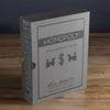 A Monopoly Vintage Bookshelf Edition box stands upright on a wooden surface, styled to look like a classic book in silver and gray with black graphics and premium components inside.