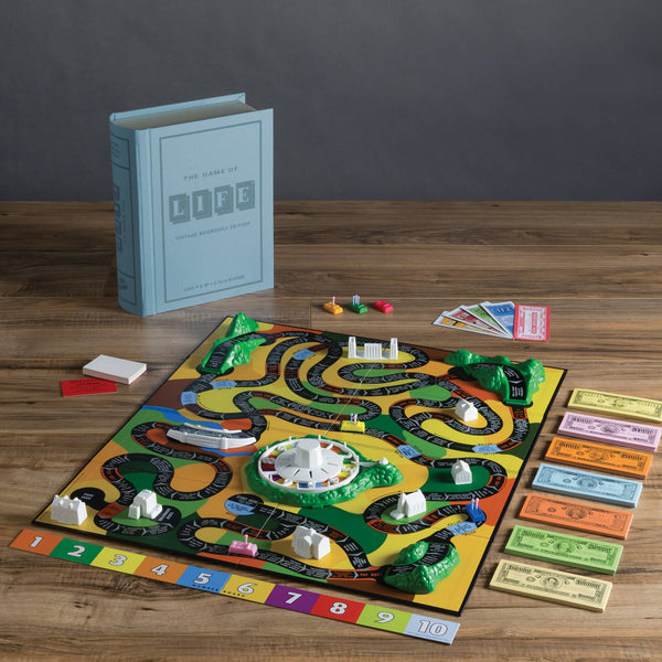 The Game of Life Vintage Bookshelf Edition is displayed on a wooden table, featuring colorful pieces, play money, cards, plastic buildings, and a book-shaped box. The board shows winding paths in a classic, elegant design.