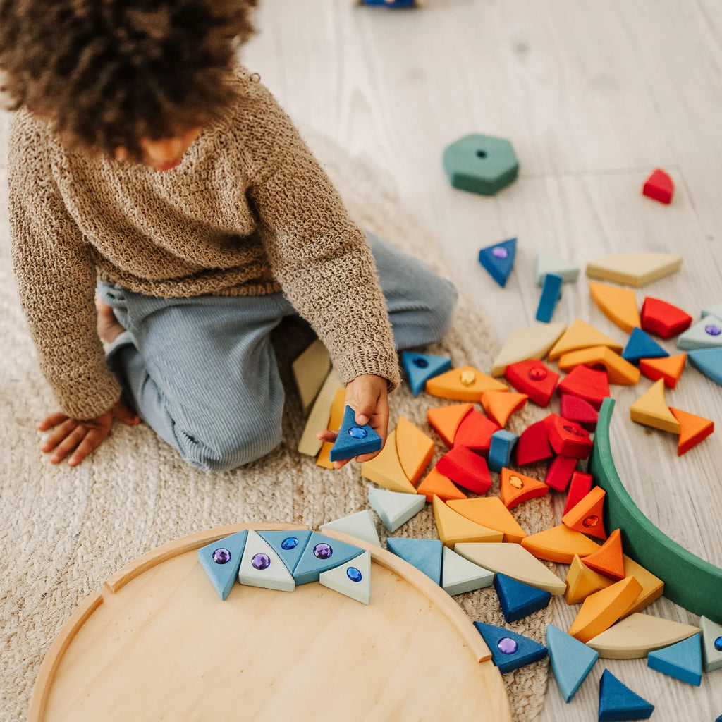 A child sits on a rug, using the Grimm’s Sparkling Mandala Sun Creative Puzzle to complete a creative design of colorful wooden triangular pieces, some inlaid with sparkling gems, on a circular wooden tray.
