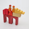 Two wooden peg people sit atop a structure made of red, orange, and yellow geometric Grimm’s Building World - Desert Sand wooden blocks.