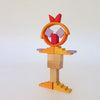 A whimsical figure from the Grimm’s Building World - Desert Sand set, built from colorful wooden blocks with a circular head, a bow, and stepped pieces for feet.