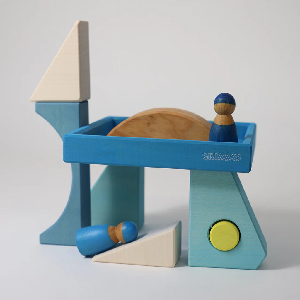A structure made from Grimm's Building Set - Blue features colorful blue, white, and natural wooden blocks plus two small blue figures—one upright and one lying down—ideal toddler toys for creative play. The "Grimm's" brand is visible on a block.