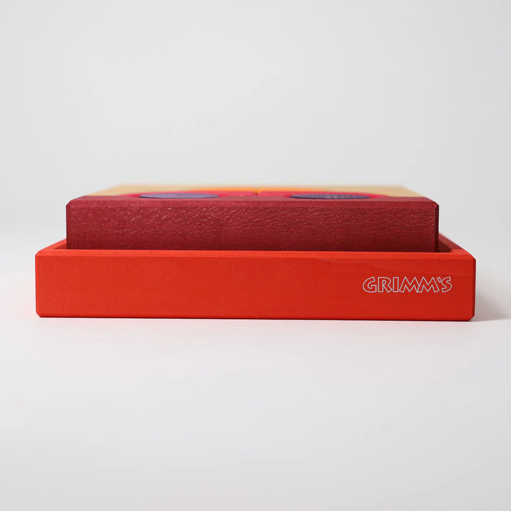 A rectangular box labeled "GRIMM'S" holds the Grimm's Building Set - Red, featuring colorful wooden toys in mostly red and orange tones, displayed against a plain white background.