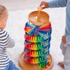 Two children play with Grimm's Assembly Set Musical Ball Run. One child drops a marble into the top spiral, watching as ramps and pegs create a vibrant rainbow pattern while the marble rolls down this educational wooden toy.