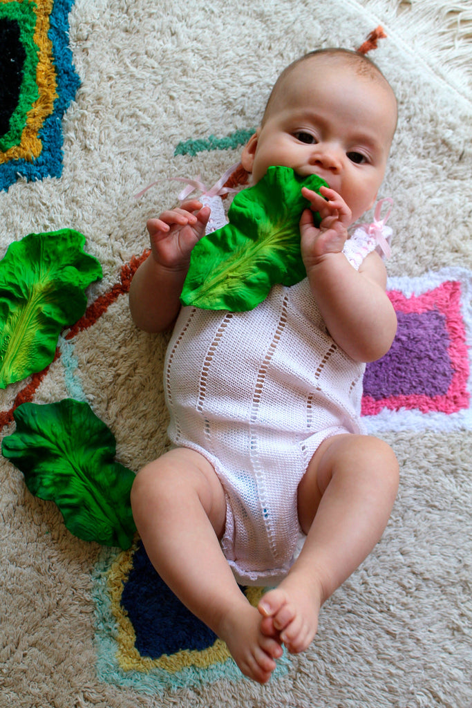 A baby in a pink knitted onesie lies on a patterned rug, holding and chewing on Kendall the Kale Teething Toy, with additional fake leaves scattered nearby.