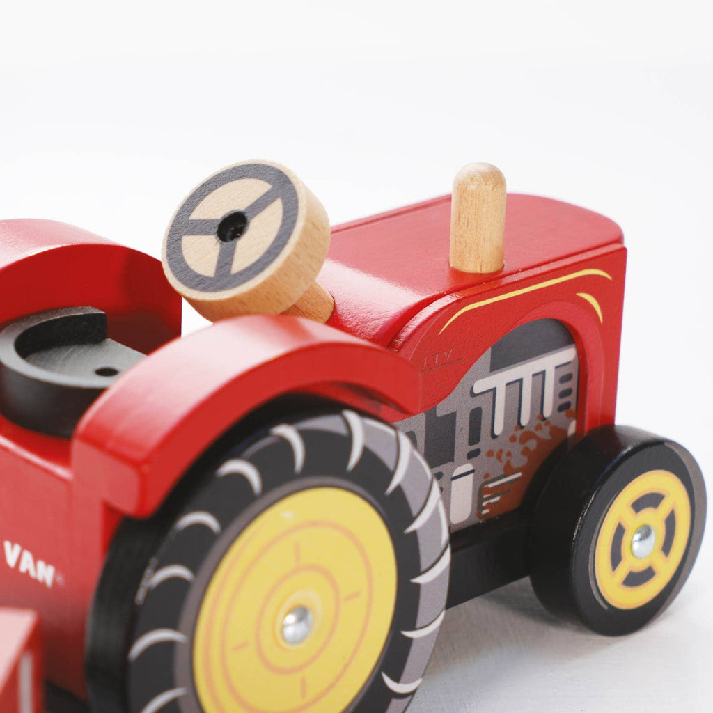 Close-up of the Farmyard Tractor & Trailer, a red wooden farm toy with black and yellow wheels, a circular steering wheel, and painted mechanical details on the side.