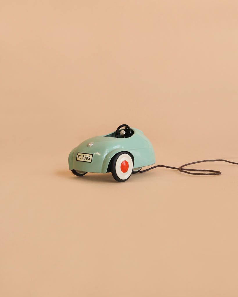 A small, mint green toy car from the Maileg Little Brother Driver Set—with white and red wheels and a black pull string—sits on a plain beige background.